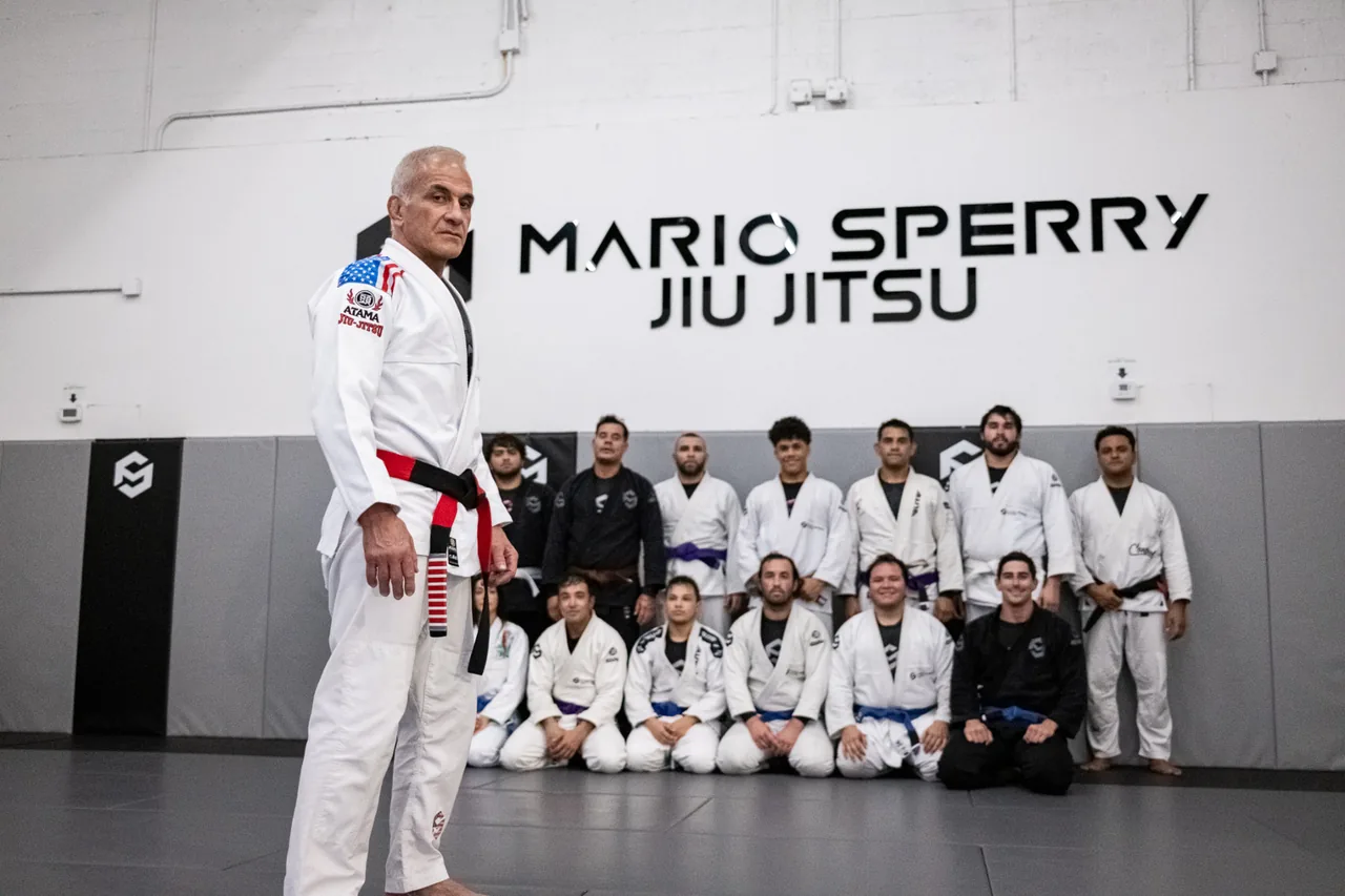 Mario Sperry wearing his coral belt, standing with students in front of the Mario Sperry Jiu-Jitsu wall at his Miami academy